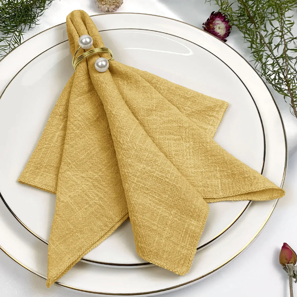 Élégante serviette de table jaune pâle, avec des perles ornementales, sur un plat blanc entouré de plantes vertes et de fleurs.
