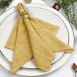 Élégante serviette de table jaune pâle, avec des perles ornementales, sur un plat blanc entouré de plantes vertes et de fleurs.