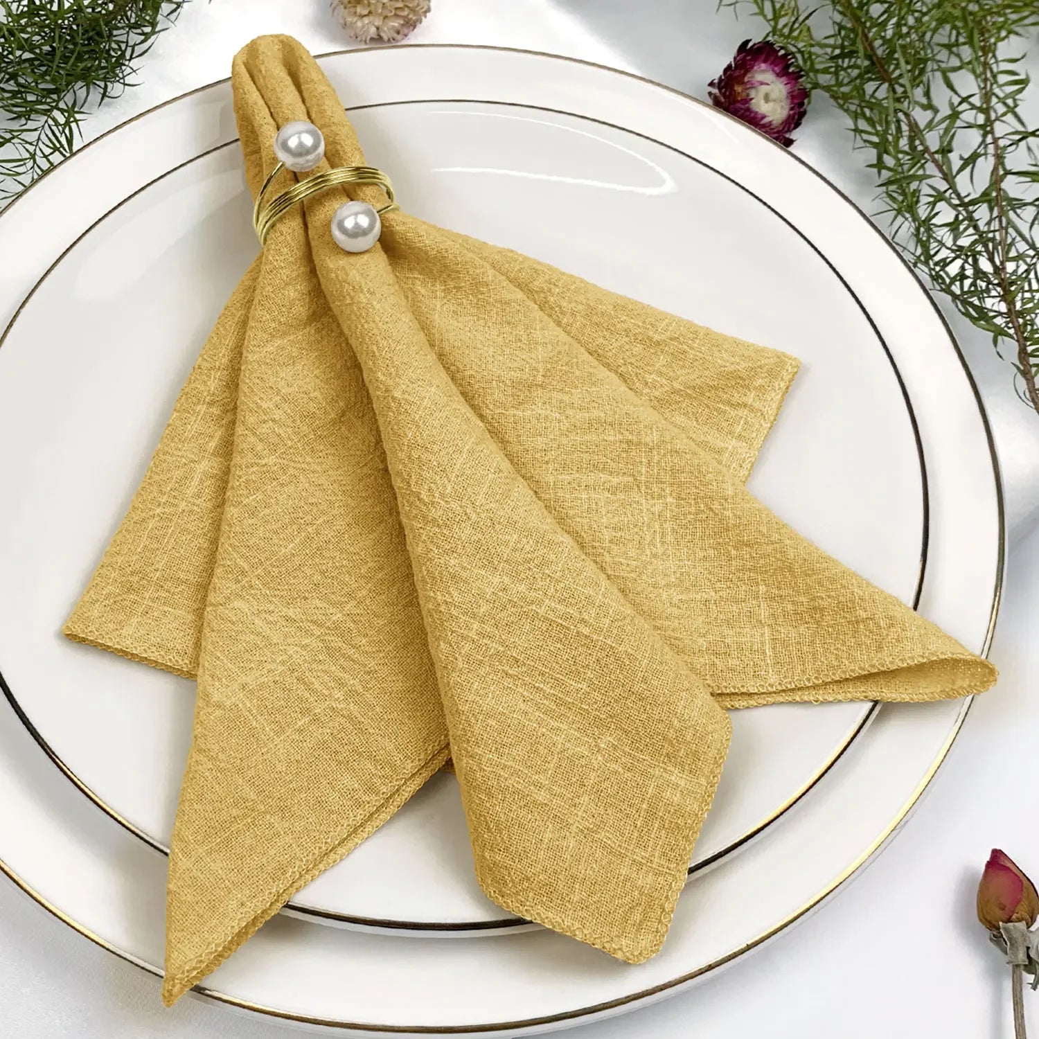 Élégante serviette de table jaune pâle, avec des perles ornementales, sur un plat blanc entouré de plantes vertes et de fleurs.