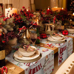 Cette image représente une table de Noël décorée de manière festive avec des bougies allumées, des branches de sapin, des baies rouges et des accessoires décoratifs, créant une ambiance chaleureuse et accueillante pour les fêtes.