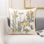 Un coussin décoratif aux motifs floraux variés avec des fleurs jaunes, blanches et bleues sur un fond blanc.