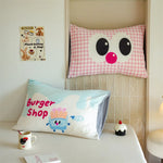 Coussin décoratif avec un visage en relief et motif vichy rose posé sur un banc, accompagné d'un autre coussin avec un personnage Burger Shop imprimé et d'un petit gâteau décoratif.
