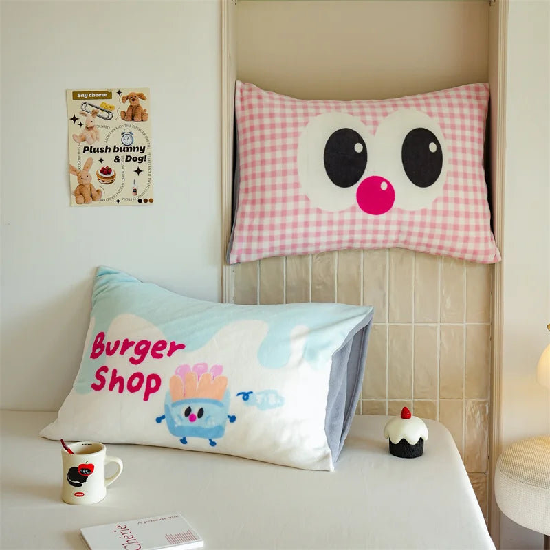 Coussin décoratif avec un visage en relief et motif vichy rose posé sur un banc, accompagné d'un autre coussin avec un personnage Burger Shop imprimé et d'un petit gâteau décoratif.