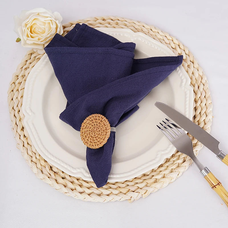 Une serviette bleue marine élégante posée sur une assiette blanche avec une nappe tressée beige et une rose blanche, le tout sur un fond blanc.