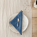 Une assiette blanche avec une cuillère en métal posée sur une serviette en lin bleu, sur un fond de bois clair.