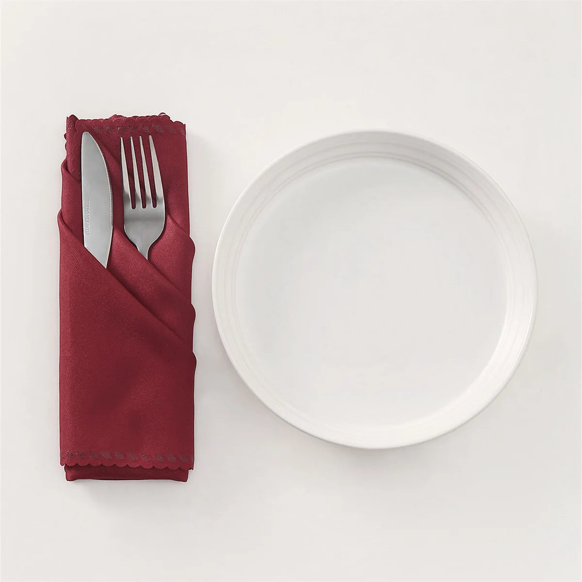 Assiette blanche vide et couverts en métal dans une serviette de table rouge sur un fond blanc.