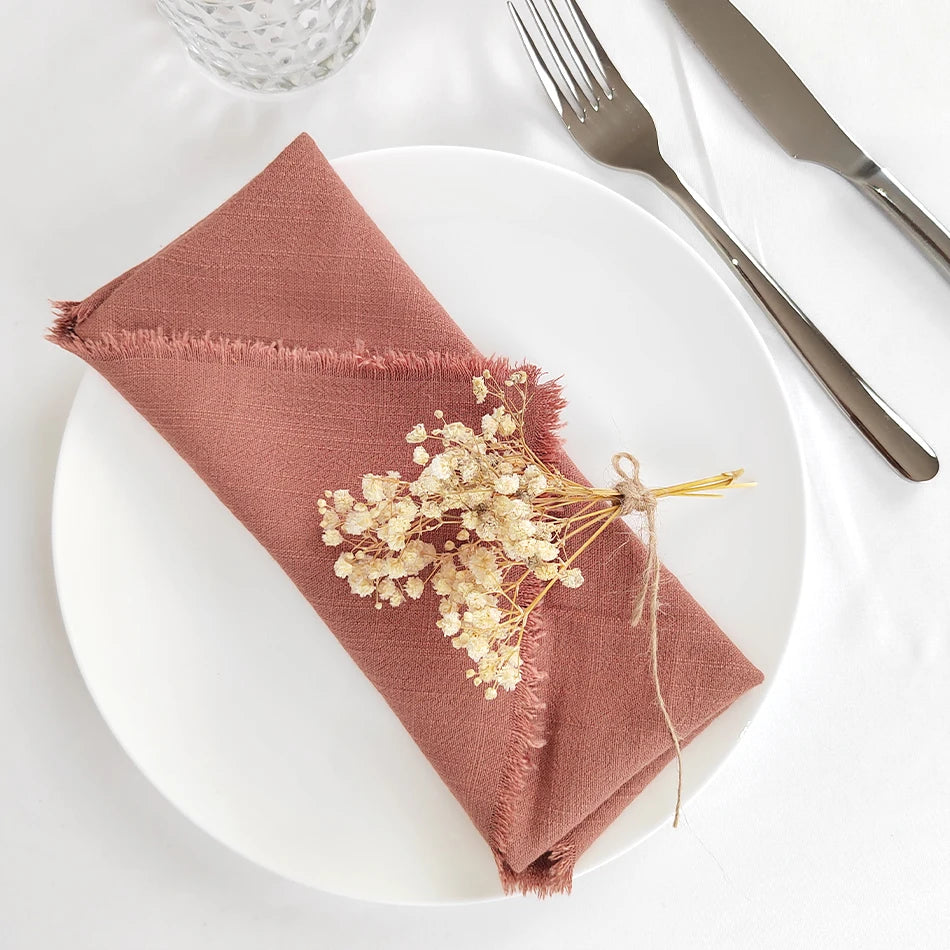 Une élégante serviette de table en couleur terracotta, avec une délicate branche de fleurs séchées nouée avec du fil, posée sur une assiette blanche, à côté d'un verre et de couverts en métal.