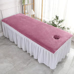Lit de massage avec couverture rose et rideau blanc, placé dans une pièce lumineuse avec des plantes vertes.