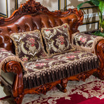 Canapé en bois taillé finement avec des ornements baroque en relief, recouvert de coussins tissu aux motifs floraux et d'un plaid frangé, créant une atmosphère luxueuse et élégante.