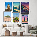 L'image présente une collection d'affiches de voyage illustrant des lieux célèbres tels que la cathédrale St Nicholas à Newcastle, la ville de Birmingham, Glasgow avec son parc d'attractions Barrowland, le village de Scalby Mills et la ville d'Alnwick, ainsi que la ville de Paris et sa tour Eiffel. Ces affiches sont disposées sur un mur blanc, encadrant un salon moderne avec un canapé blanc, une table basse en bois et un fauteuil gris.