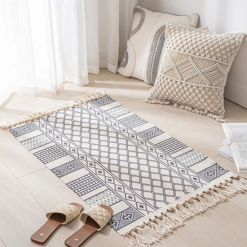 Un tapis décoratif en coton avec des motifs géométriques en noir et blanc se trouve sur le sol d'une pièce lumineuse, accompagné de coussins et d'un rideau blanc.