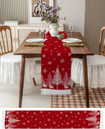 Une table joliment dressée avec des assiettes, des verres et une décoration de Noël, notamment un chemin de table rouge orné de motifs de flocons de neige et de silhouettes de cerfs.
