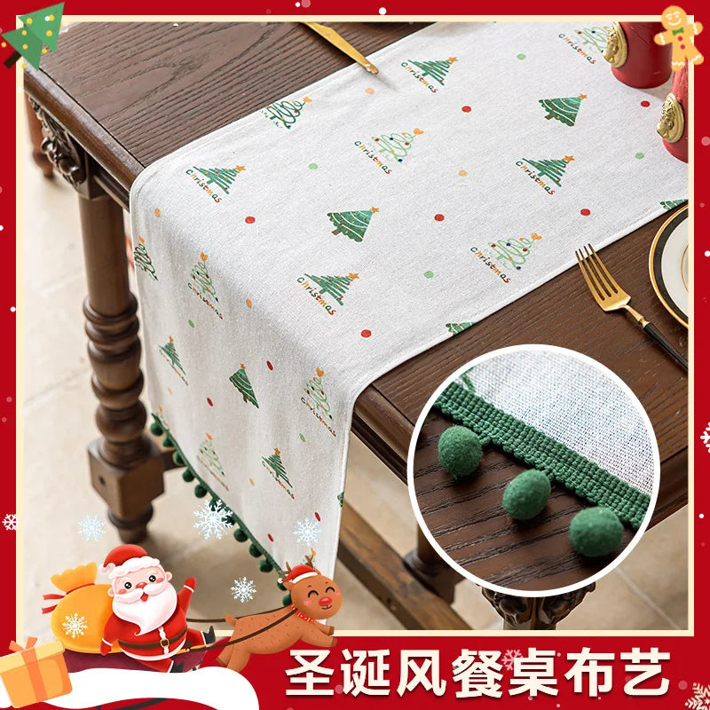 Cette image montre un ensemble de table avec une nappe à motifs d'arbres de Noël et de boules de Noël vertes. On voit également des décorations de Noël telles que le Père Noël et un renne.