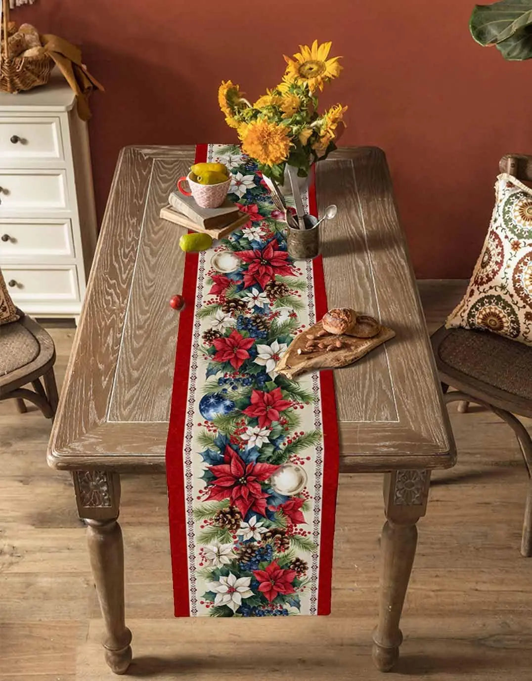 Une table en bois rustique ornée d'un chemin de table festif aux motifs de fleurs, de baies et de pommes de pin, agrémentée d'un bouquet de tournesols et d'autres éléments décoratifs.