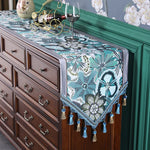 Cette image présente un meuble en bois foncé recouvert d'un chemin de table décoratif aux motifs floraux dans des tons de vert et bleu, accompagné de verres à vin et d'objets de décoration.