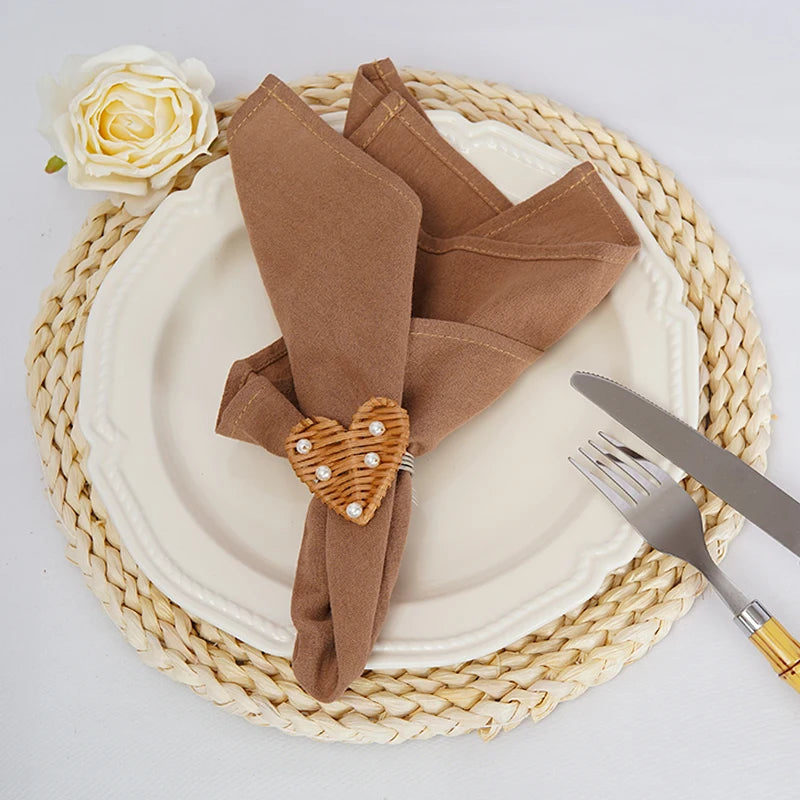 L'image montre un couvert avec une serviette de table marron pliée avec un motif en forme de cœur en perles sur la table, décorée d'une rose blanche et placée sur un tapis tressé beige.