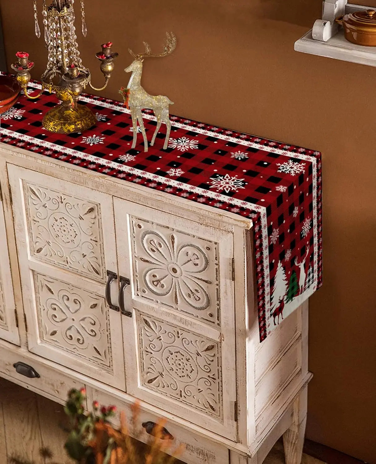 Meuble vintage décoré de motifs floraux et de figurines de renne doré sur un chemin de table aux motifs de flocons de neige rouges et noirs, créant une atmosphère chaleureuse et festive pour la période des fêtes.