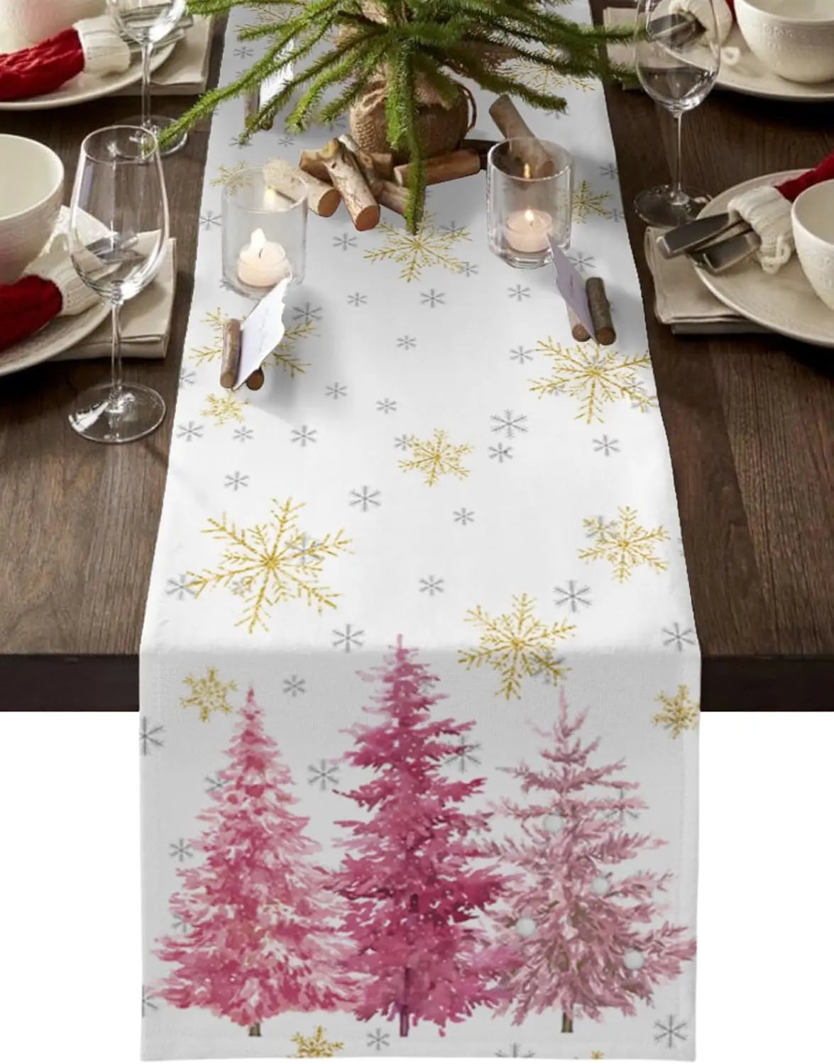 Une table de fête joliment décorée avec une nappe blanche imprimée de flocons de neige dorés et de sapins roses, entourée de verres à vin et de bougies.