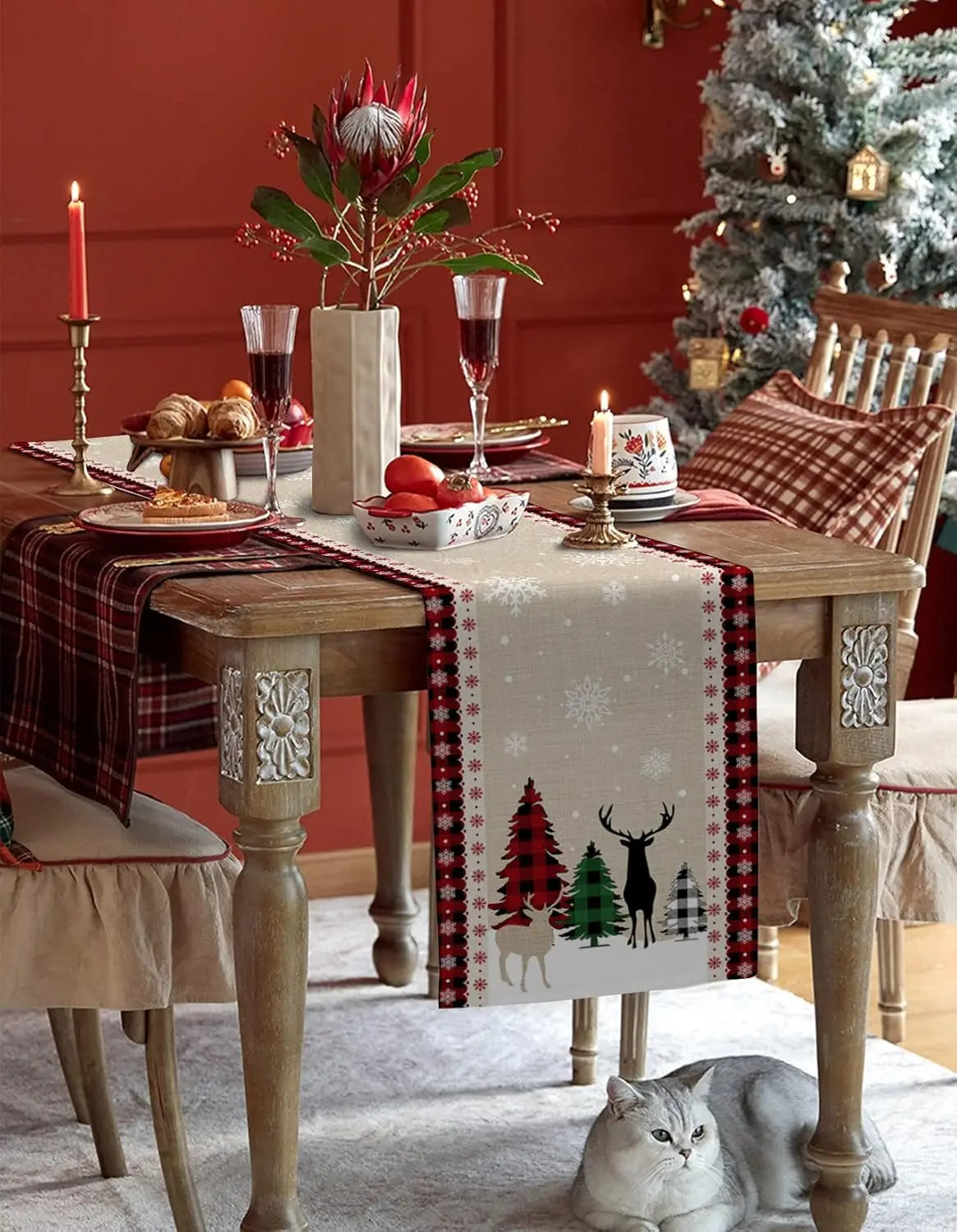 Une table de Noël festive avec une nappe à motifs de flocons de neige et de cerfs, des bougies, des fleurs et des décorations sur un fond rouge chaleureux.