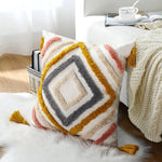 Coussin décoratif multicolore en peluche avec des franges et des glands, sur un lit douillet avec une couverture et un panier en osier.