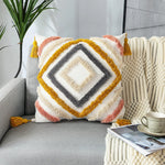 Coussin décoratif aux motifs géométriques aux couleurs chaudes et textures douces, placé sur un canapé gris dans un intérieur moderne avec plante verte.