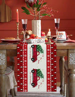 Cette image montre une table de fête de Noël décorée avec des fleurs, des bougies, des verres à vin et un chemin de table en tissu rouge avec des motifs de voitures vintage et de flocons de neige, créant une ambiance festive et chaleureuse.