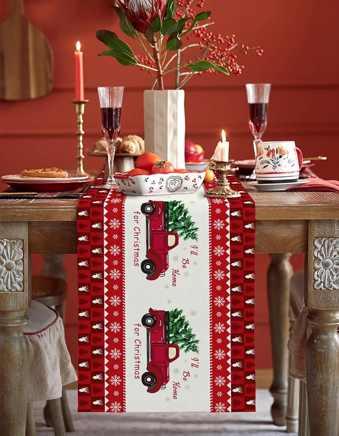Cette image montre une table de fête de Noël décorée avec des fleurs, des bougies, des verres à vin et un chemin de table en tissu rouge avec des motifs de voitures vintage et de flocons de neige, créant une ambiance festive et chaleureuse.