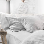 Une chambre douillette avec des draps de lin blancs, une tête de lit en tricot et un petit tabouret en bois, créant une atmosphère reposante et confortable.
