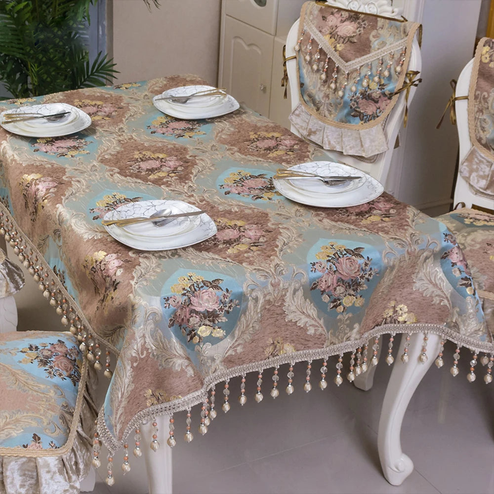 Une nappe élégante et somptueuse, richement décorée de motifs floraux aux tons chauds et doux, ornée de pompons et de franges, posée sur une table avec des assiettes et des couverts.