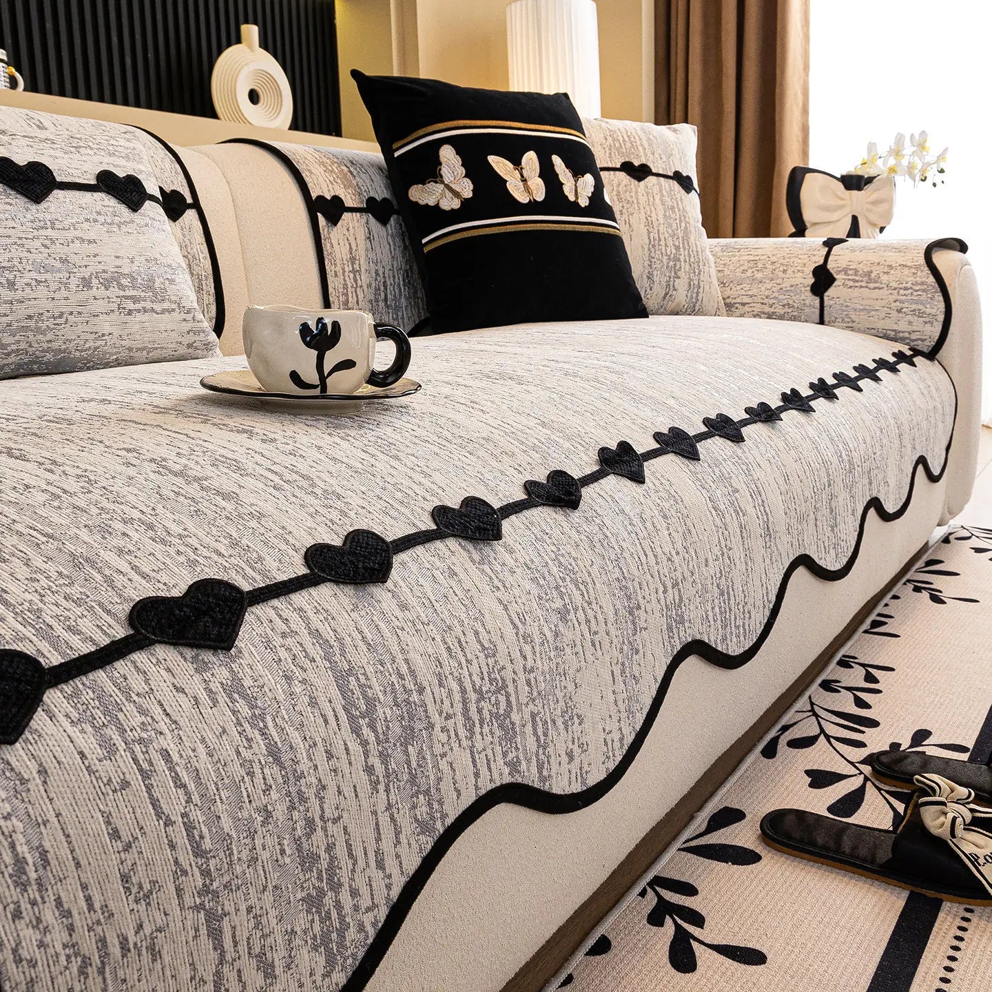 Lit confortable avec linge de lit décoré de motifs de cœurs et de papillons noirs sur fond gris clair, orné d'un coussin assorti et d'une tasse design sur la table basse.