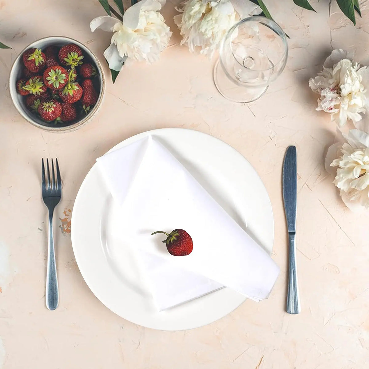 Composition élégante avec assiette blanche, couverts, fraises fraîches et fleurs de pivoines blanches sur fond clair.