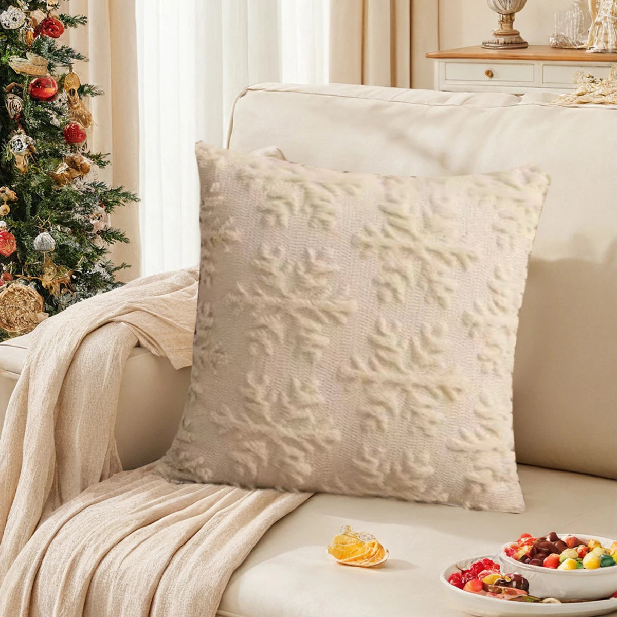 Gros plan sur un oreiller décoratif en peluche blanche sur un canapé beige, avec un arbre de Noël orné dans le fond et un bol de fruits sur une table basse.