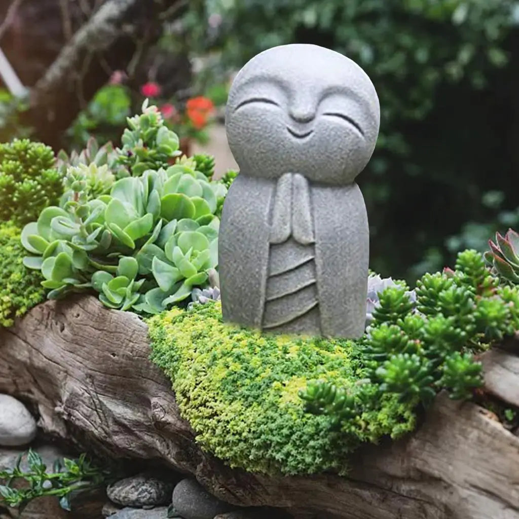 statue personnage yeux fermés sourire gris clair texture granuleuse plantes succulentes mousse fleurs zen