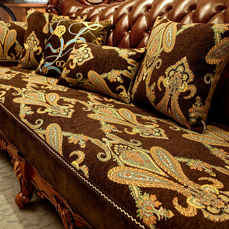 Des coussins décoratifs en velours marron avec un motif floral doré et des détails ethniques ornent un canapé en cuir brun, créant une ambiance chaleureuse et élégante dans cette pièce.