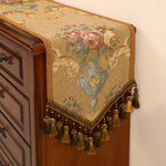 Un élégant chemin de table en tissu orné de fleurs épanouies et de motifs floraux, entouré d'une frange en dorée subtile, confère une touche de sophistication vintage à ce meuble en bois.