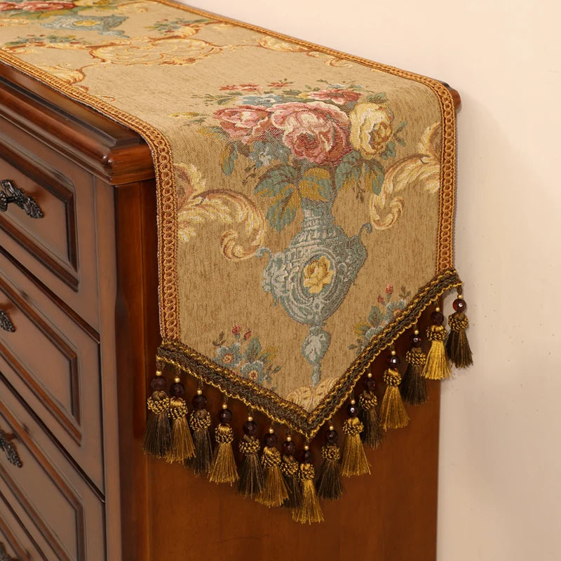 Un élégant chemin de table en tissu orné de fleurs épanouies et de motifs floraux, entouré d'une frange en dorée subtile, confère une touche de sophistication vintage à ce meuble en bois.
