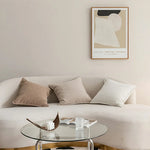 Ce salon minimaliste comprend un canapé beige confortable orné de coussins assortis, une table basse ronde en verre et une œuvre d'art abstraite accrocheée au mur.