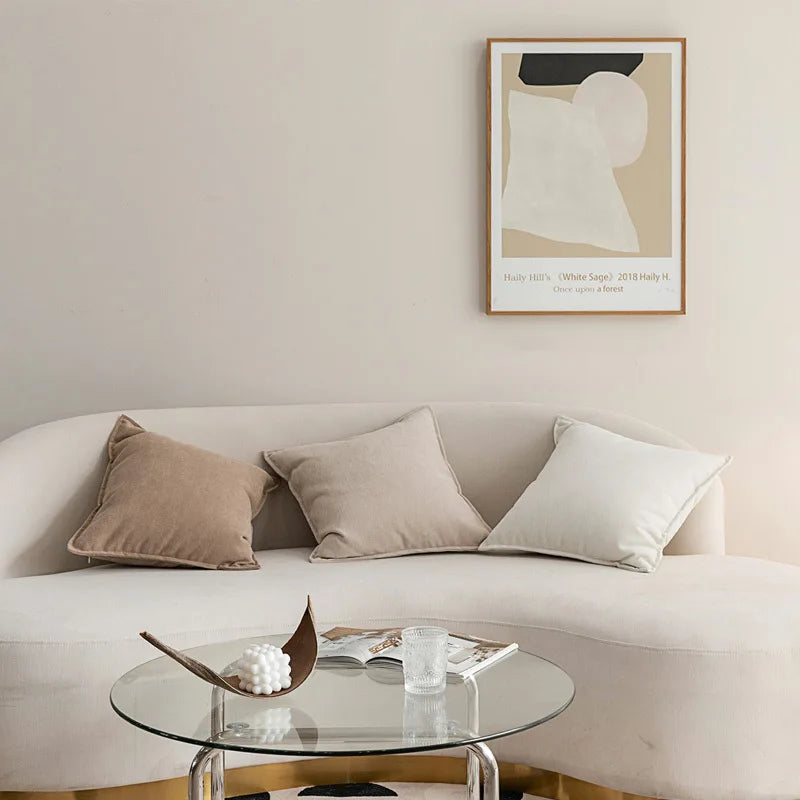 Ce salon minimaliste comprend un canapé beige confortable orné de coussins assortis, une table basse ronde en verre et une œuvre d'art abstraite accrocheée au mur.