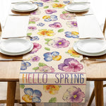 Une nappe de table colorée avec de beaux motifs de fleurs de printemps dans des tons de violet, jaune et bleu, complétée par le texte "Hello Spring" pour célébrer l'arrivée de la saison printanière.