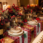Cette image montre une belle table de Noël décorée avec une composition florale de brindilles de sapin, de baies rouges et de bougies allumées, ainsi que des cadeaux emballés dans du papier cadeau rouge et blanc festif.