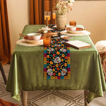 Cette image montre une décoration de table avec une nappe verte, une chemin de table coloré orné de motifs de crânes mexicains et de fleurs, ainsi que différents plats et éléments de vaisselle posés sur la table.