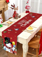 Table de Noël ornée de figurines décoratives et d'une nappe imprimée d'un motif de Noël avec texte en anglais "Merry Christmas". L'ensemble crée une atmosphère festive et chaleureuse pour les fêtes de fin d'année.