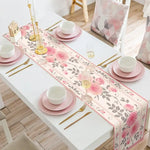 Une nappe de table blanche avec des motifs floraux rose et gris, accompagnée de vaisselle rose et de couverts dorés, créant une ambiance chic et raffinée.