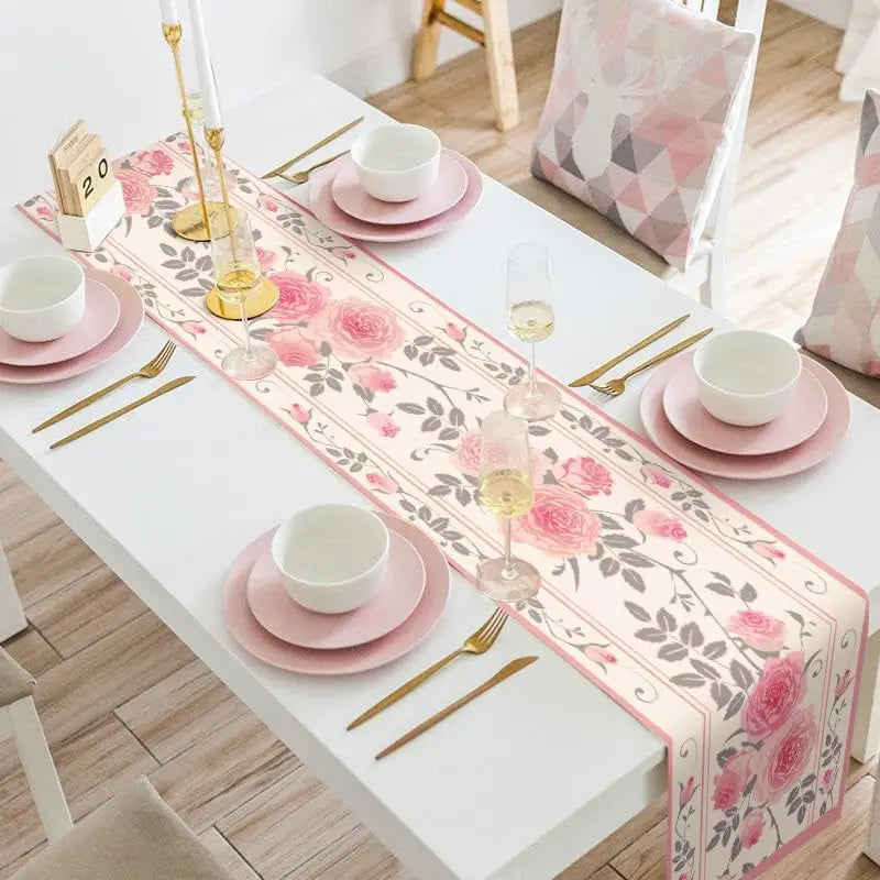 Une nappe de table blanche avec des motifs floraux rose et gris, accompagnée de vaisselle rose et de couverts dorés, créant une ambiance chic et raffinée.