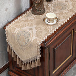 Sur un meuble en bois sombre, un chemin de table en dentelle beige orné de motifs floraux délicats et de franges apporte une touche élégante à cette scène vintage.