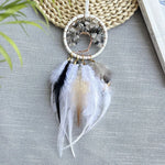 Un joli attrape-rêves fait main avec des plumes blanches et grises, un cadre en fil de perles et des cristaux bruts, posé sur un fond de feuillage vert.