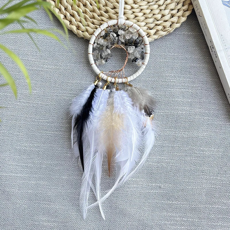 Un joli attrape-rêves fait main avec des plumes blanches et grises, un cadre en fil de perles et des cristaux bruts, posé sur un fond de feuillage vert.