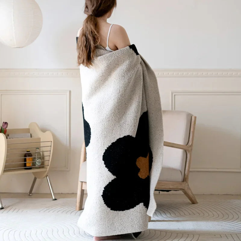 Une femme aux cheveux longs enveloppée dans une couverture en peluche blanche et noire, debout dans une pièce lumineuse.