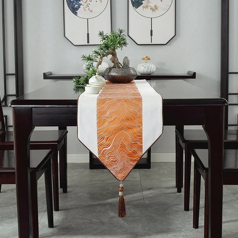 Cette image montre une table à manger élégante avec un chemin de table orange décoré de motifs vagues, un arrangement de plantes et de céramiques sur le centre de la table, et des chaises en bois sombre autour.