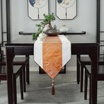 Cette image montre une table à manger élégante avec un chemin de table orange décoré de motifs vagues, un arrangement de plantes et de céramiques sur le centre de la table, et des chaises en bois sombre autour.